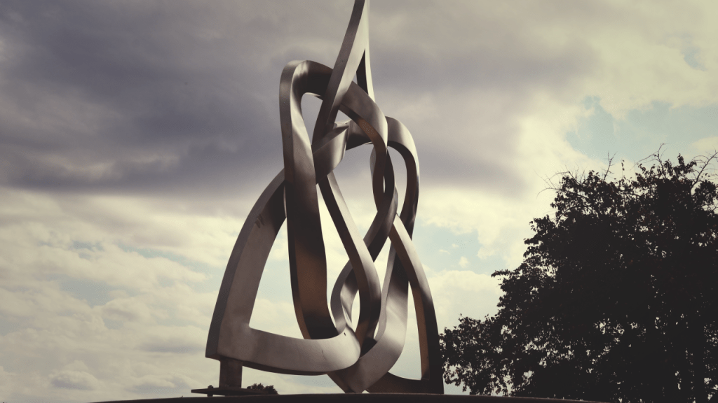 A metal sculpture depicting a Celtic trinity shape is intertwined with a metal heart. The sculpture, titled 'Eternal Heart' is in angled and shown against a cloudy sky and a tree in the background.